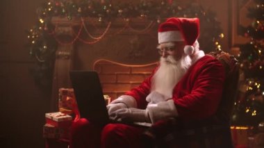 An elderly man in a Santa Claus costume sits in a beam of light at home against the background of a fireplace and a Christmas tree on Christmas eve and types on a laptop while sitting on the Internet.