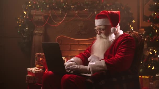An elderly man in a Santa Claus costume sits in a beam of light at home against the background of a fireplace and a Christmas tree on Christmas eve and types on a laptop while sitting on the Internet. — 비디오