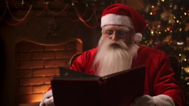 a man with a white beard in a Santa Claus suit sitting in a chair against the background of a decorated Christmas tree with a garland reads and flips through the pages of a book