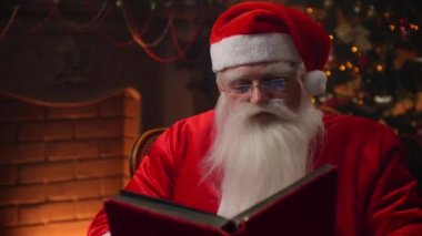 An elderly man with a white beard in a Santa Claus suit sitting in a chair against the background of a decorated Christmas tree with a garland reads and flips through the pages of a book