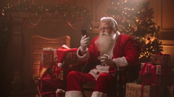 An elderly man with a white beard listens to music in a Santa Claus costume on Christmas eve. Santa Claus in the new year — Stok video