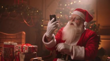 Santa Claus uses a smartphone and talks via video link while conducting a conference. Santa Claus and selfie camera, online broadcasts.