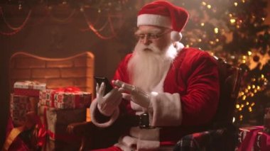 Santa Claus is sitting in a chair on the background of a Christmas tree using a smartphone and chatting on the Internet and reading messages from children.