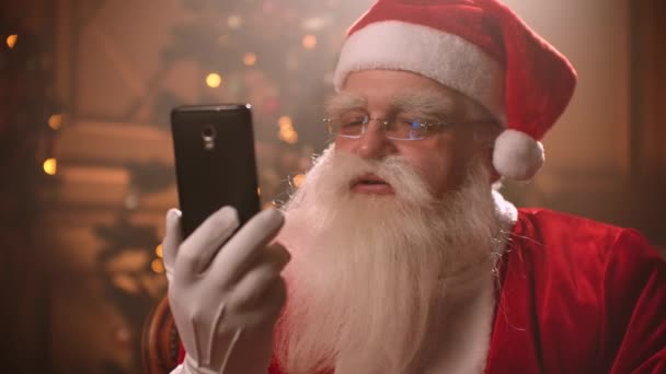 Santa Claus utiliza un teléfono inteligente y habla a través de un enlace de vídeo mientras conduce una conferencia. Santa Claus y cámara selfie, transmisiones en línea . — Vídeo de stock