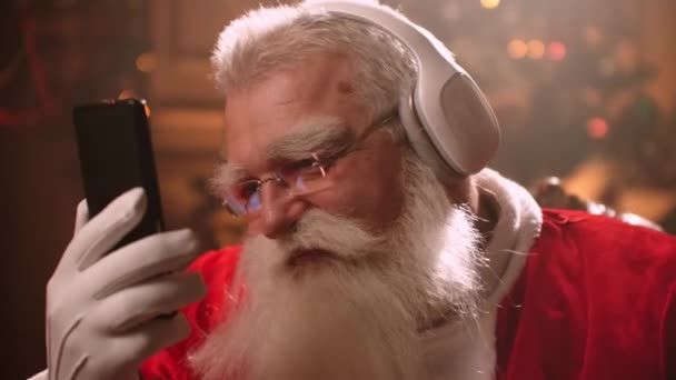 Пожилой человек с белой бородой слушает музыку в костюме Санта-Клауса в канун Рождества. Санта Клаус в новом году — стоковое видео