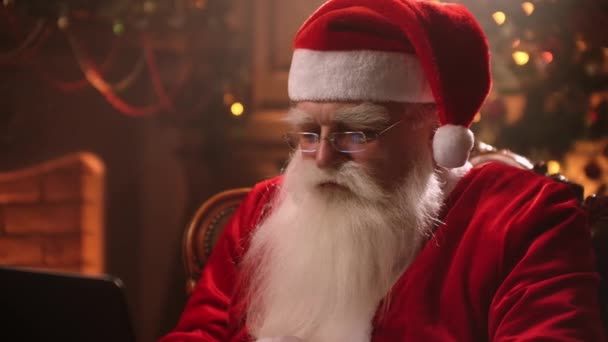 De Kerstman met een witte baard zit in een stoel en houdt zich bezig met internetbankieren. hun rekeningen en e-mail bekijken. — Stockvideo