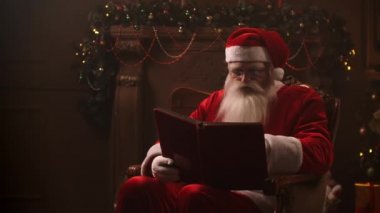 A beautiful Santa Claus sitting in a chair on the background of a decorated Christmas tree with a garland reads and flips through the pages of a book