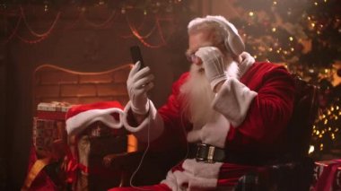 Senior man with a white beard listens to music with headphones and a Santa Claus costume on Christmas eve. Santa Claus in the new year. Jolly senior man dancing to music