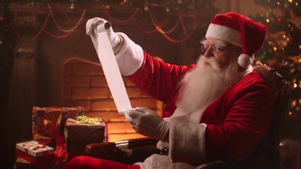 Санта Клаус з бородою і костюмом сидить на стільці і читає сувій зі списком дітей на подарунки. — стокове відео