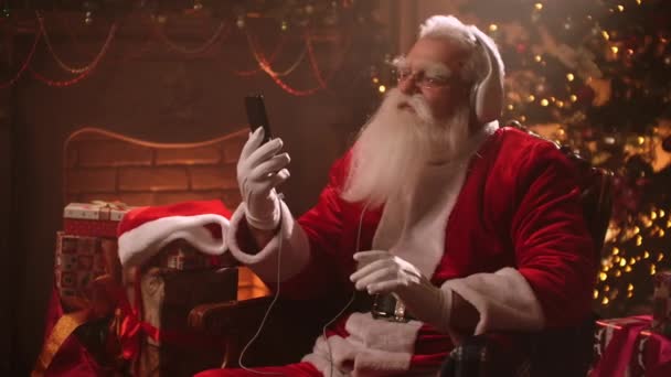 Senior man with a white beard listens to music with headphones and a Santa Claus costume on Christmas eve. Santa Claus in the new year. Jolly senior man dancing to music — Stok video