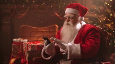 Santa Claus is sitting in a chair on the background of a Christmas tree using a smartphone and viewing his online Bank accounts.