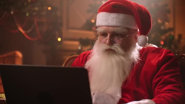 In a magical atmosphere Santa Claus uses a laptop to work and distribute gifts to children against the background of a Christmas tree and a fireplace — Stock Video