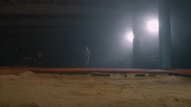 A sporty woman runs up in a stadium and performs a long jump in slow motion in a stadium on a dark background — 图库视频影像