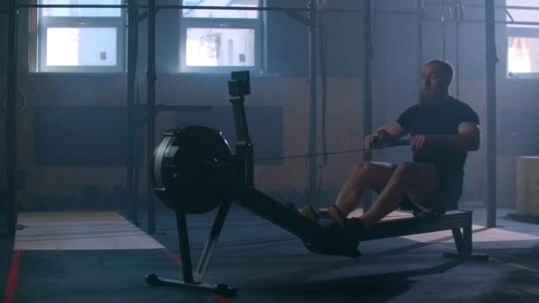Un hombre fuerte en cámara lenta tira de una cuerda en un simulador de remo. Entrenamiento cardiovascular para un hombre en un gimnasio atmosférico . — Vídeos de Stock