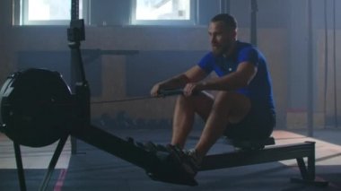 Ağır çekimde güçlü bir adam kürek simülatöründe ipi çeker. Atmosferik fitness odasında bir adam için kardiyo eğitimi.