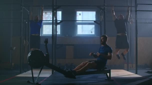 Tres hombres trabajan juntos en una sala de fitness. Un hombre tira de una máquina de remo, y dos realizan pull-UPS en la barra. Entrenamiento cruzado en cámara lenta . — Vídeo de stock