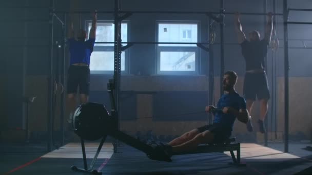 Tres hombres trabajan juntos en una sala de fitness. Un hombre tira de una máquina de remo, y dos realizan pull-UPS en la barra. Entrenamiento cruzado en cámara lenta . — Vídeos de Stock