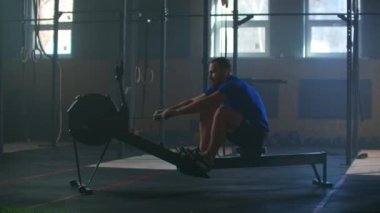 Ağır çekimde güçlü bir adam kürek simülatöründe ipi çeker. Atmosferik fitness odasında bir adam için kardiyo eğitimi.