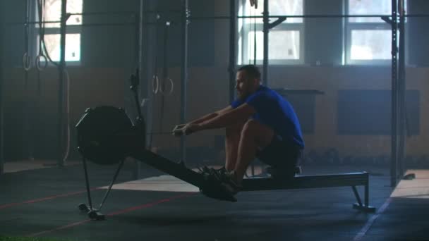Un hombre fuerte en cámara lenta tira de una cuerda en un simulador de remo. Entrenamiento cardiovascular para un hombre en un gimnasio atmosférico . — Vídeo de stock