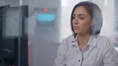 Beyaz önlüklü bir kadın doktor cam duvarın arkasındaki parlak ofiste oturan monitör ekranına bakar. Modern klinik.