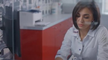Laboratuvar önlüğü giymiş profesyonel bir kadın doktor, ofiste cam bir duvarın arkasında monitörle bilgisayarda yazıyor..