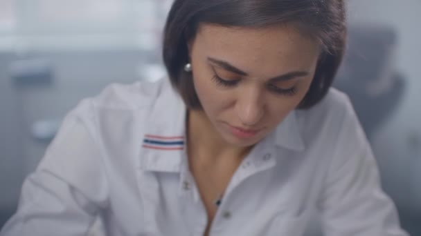 A female doctor sits at a table and writes with a pen writing out a prescription to a patient, behind a glass wall. Writing with a pen close-up — Stock video