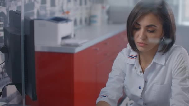 Laboratuvar önlüğü giymiş profesyonel bir kadın doktor, ofiste cam bir duvarın arkasında monitörle bilgisayarda yazıyor.. — Stok video