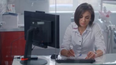 Laboratuvar önlüğü giymiş profesyonel bir kadın doktor, ofiste cam bir duvarın arkasında monitörle bilgisayarda yazıyor..
