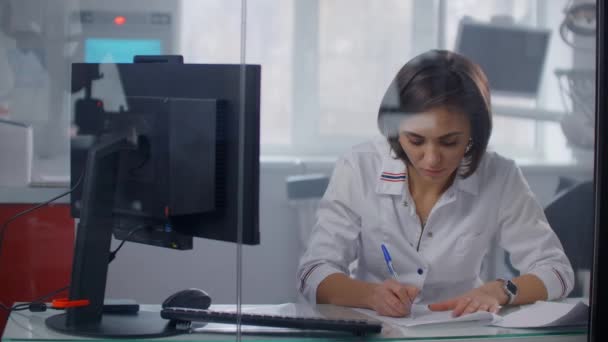 A female doctor sits at a table and writes with a pen writing out a prescription to a patient, behind a glass wall. Writing with a pen close-up — Stock video