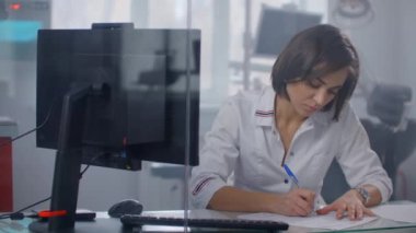 Kadın bir doktor masaya oturur ve hasta çizelgesinde bir kalemle yazar, cam bir duvarın arkasında. Bir kalem yakın plan ile yazılıyor