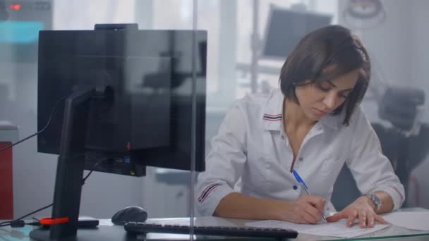 Una doctora se sienta en una mesa y escribe con un bolígrafo en la tabla de pacientes, detrás de una pared de vidrio. Escribir con una pluma de cerca — Vídeos de Stock