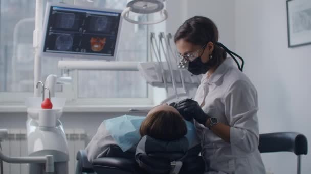 Une jeune dentiste en jumelles et un manteau blanc commencent à examiner et à traiter une patiente. Traitement dentaire et installation d'appareils dentaires — Video
