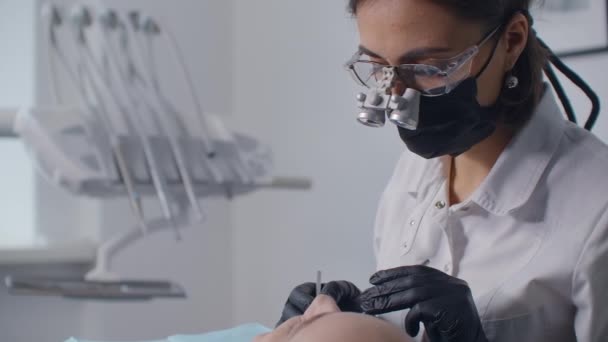 Una dentista donna in binocolo e una maschera esaminano la cavità orale di una paziente sdraiata su una sedia. Il concetto di un sorriso sano — Video Stock