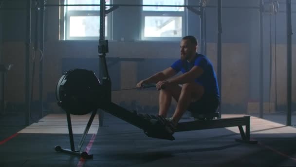 Cámara lenta: un hombre en una sala de fitness atmosférica al sol en una máquina de remo — Vídeo de stock