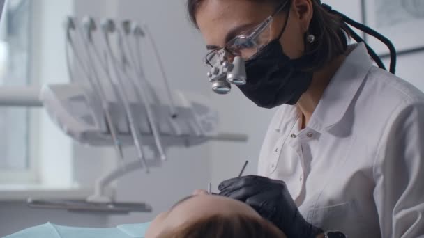 Mladá zubařka v dalekohledu a bílý kabát začnou vyšetřovat a léčit pacientku. Zubní ošetření a montáž rovnátek — Stock video