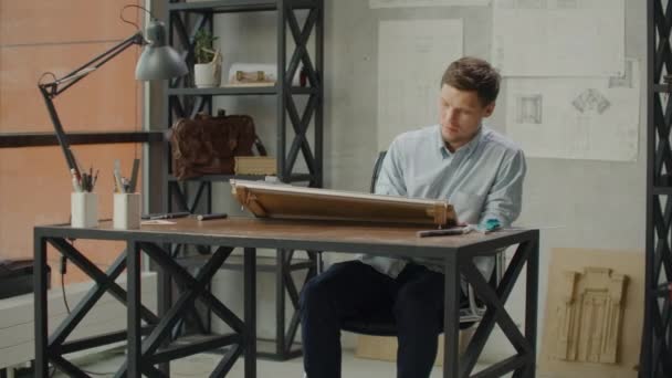 Engineer works in a bright office with a large window, concentrates and draws blueprints. Workplace of an architect or designer: loft style, minimalistic interior, drawings. — Stock Video