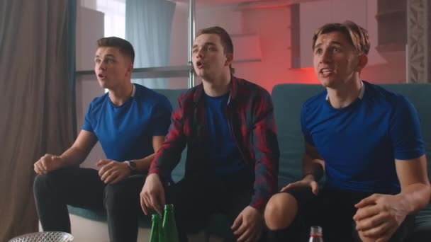 Група чоловіків у блакитних футболках. Дивитися футбольний матч з друзями, сидячи на дивані, підбадьорювати і аплодувати, дивлячись прямо на камеру — стокове відео