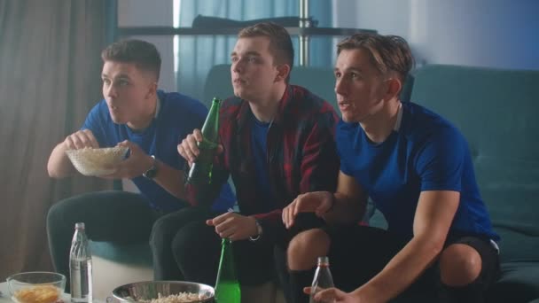 Drei Männer, die mit einem Bier auf dem Sofa sitzen und Fernsehen mit einer Sportübertragung sehen, springen und jubeln zusammen und umarmen sich — Stockvideo