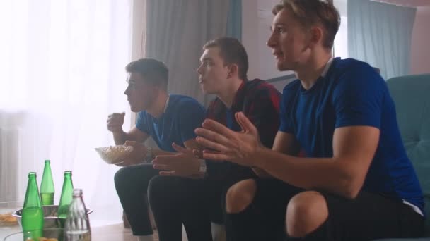 青いtシャツの3人の男性ソファに座っている友人とサッカーの試合を見て、応援し、カメラを直接見て拍手 — ストック動画