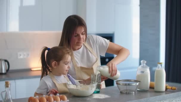 Kız ve anne birlikte yemek pişiriyor. Kadın, yoğurmak için hamura süt döken ellerdir. Annesi küçük bir kızın pasta pişirmeyi öğrendiği bir asistan. Okul öncesi yaşı. — Stok video