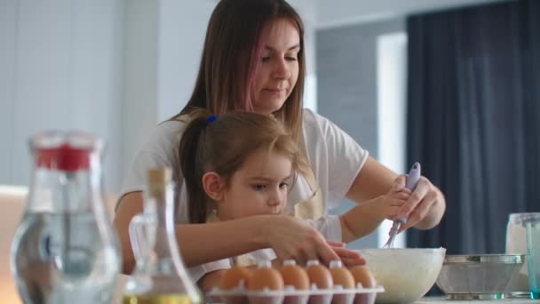 Una madre sonriente ayuda a su hija a aprender a hacer un pastel. Enseñar a una niña pequeña ayuda en casa . — Vídeo de stock