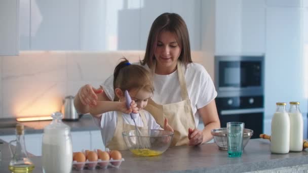 Mamá le enseña a su hija a batir la masa con un batidor para hacer un pastel. Madre amorosa e hija cocinan un pastel juntas. Mamá es asistente de delantal chica — Vídeos de Stock