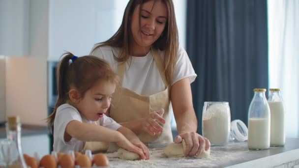 Mother teaching daughter to knead dough for cookies on kitchen in slow motion. Mom teaching kid daughter learning kneading dough with rolling pin, funny child girl helping mother preparing cookies. — Stock Video