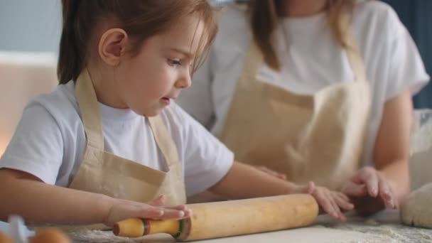 Любляча щаслива мама навчає милу доньку вчитися замішувати тісто з прокатним штифтом, смішна дівчинка, яка допомагає мамі готувати печиво для випічки, граючи на сучасній кухні вдома — стокове відео