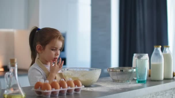 Küçük bir kız annesiyle hamur pişirdikten sonra mutfakta önlükle parmağını yalıyor. — Stok video