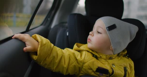 Powolny ruch: Młody wymarzony chłopiec w żółtej kurtce siedzi na foteliku dziecięcym z tyłu samochodu i patrzy przez okno uśmiechnięty. Marzycielski, troskliwy chłopiec od 2 lat. Emocje twarzy małego podróżnika. — Wideo stockowe