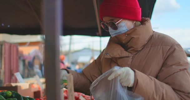 En kvinna i mask och handskar köper grönsaker på marknaden i karantänläge. Coronavirus köper grönsaker och frukt. Inköp av produkter under en pandemi — Stockvideo