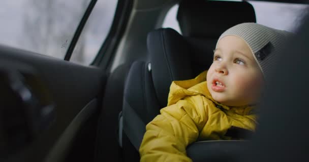 Mouvement lent : Un jeune garçon rêveur vêtu d'une veste jaune s'assoit dans un siège enfant à l'arrière d'une voiture et regarde par la fenêtre en souriant. Garçon rêveur et réfléchi de 2 ans. Émotions faciales du petit voyageur . — Video