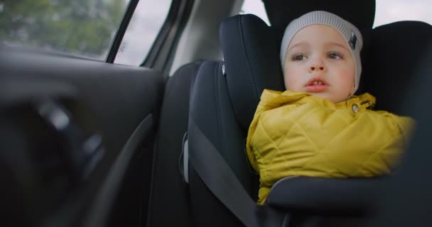 Медленное движение: молодой мечтательный мальчик в желтой куртке сидит на детском сиденье на заднем сиденье автомобиля и смотрит в окно улыбаясь. Задумчивый 2-летний мальчик. Лицевые эмоции маленького путешественника . — стоковое видео