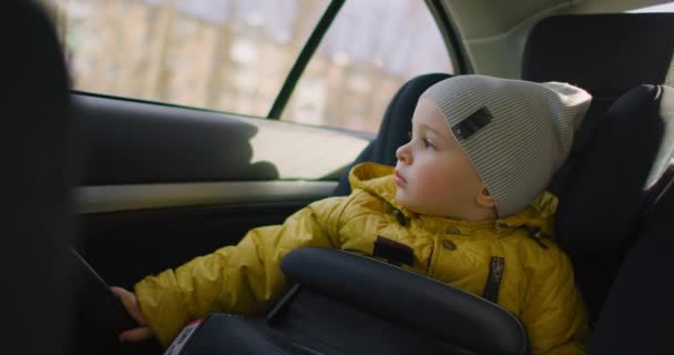 Movimiento lento: Un niño de ensueño con una chaqueta amarilla se sienta en un asiento de niño en la parte trasera de un coche y mira por la ventana sonriendo. Soñador niño pensativo de 2 años. Emociones faciales de viajero pequeño . — Vídeos de Stock